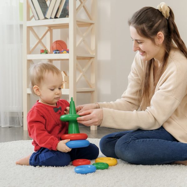 Young loving mother helping her baby son assembling colorful toy tower or pyramid.